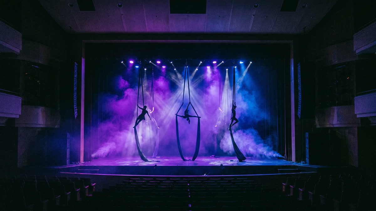 A dramatic wide shot of a theatrical stage bathed in coloured spotlights with silhouettes of acrobats mid-performance